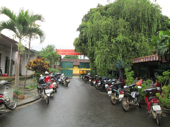 Cổng vào Di tích lịch sử Nhà đày Buôn Ma Thuột trên đường Tán Thuật bị bao vây, che lấp bởi cây cối và xe cộ của khách uống  cà phê.