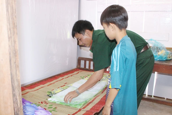 Cán bộ Đồn Biên phòng Cửa khẩu Đắk Ruê hướng dẫn con nuôi - cháu Phạm Vũ Đình Hiếu cách gấp chăn màn.